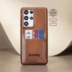 以下が含まれることがあります： 茶色の革製携帯電話ケース。カードホルダーと「Jeremy」という名前が刻印されています。ケースにはカメラの切り抜きがあり、「Wireless Charging Compatible」と「Genuine 100% Leather」の文字があります。ケースには2枚のカードが入っています。