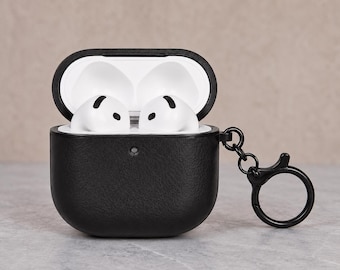 May include: A black protective case for wireless earbuds, featuring a textured finish and an attached black circular keychain ring. The case is open, revealing two white wireless earbuds inside, with a white interior lining visible at the top.
