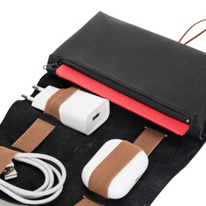 Puede incluir: Un rollo de viaje de cuero negro con correas el&aacute;sticas marrones para sujetar un cargador de tel&eacute;fono blanco, una funda de auriculares inal&aacute;mbricos blanca y un cable de carga blanco.