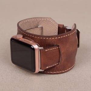 May include: A brown leather watch band with a rose gold Apple Watch attached. The band is thick, with white stitching and a buckle closure. The watch face is black.