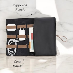 Puede incluir: Organizador de viaje de cuero negro con una bolsa con cremallera y bandas elásticas para cables para guardar cables y accesorios. El organizador está abierto para revelar el interior con bandas elásticas marrones y un cable blanco.