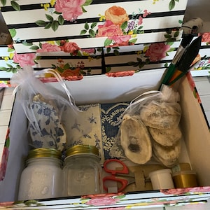 May include: A white box with a floral pattern on the lid and sides. The box is open and contains two glass jars with gold lids, a pair of scissors, a small bag of white beads, a roll of gold ribbon, and a few seashells.