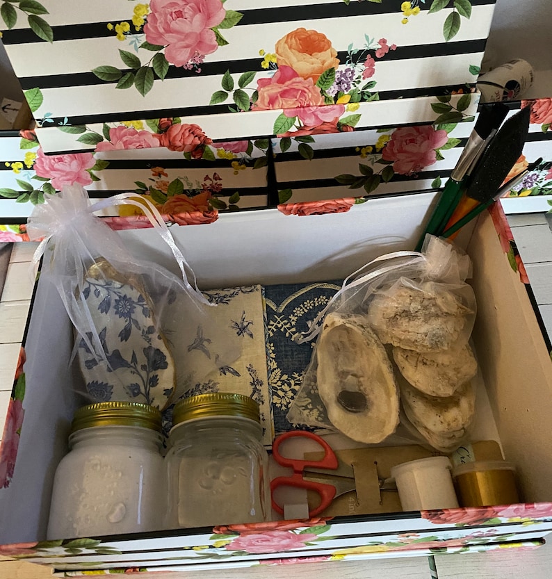 May include: A white box with a floral pattern on the lid and sides. The box is open and contains two glass jars with gold lids, a pair of scissors, a small bag of white beads, a roll of gold ribbon, and a few seashells.