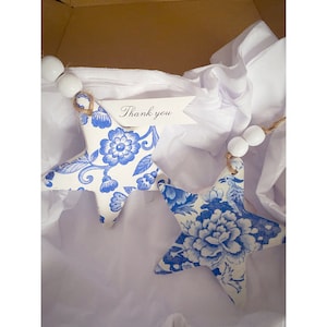 May include: Two blue and white star-shaped ornaments with floral patterns. The ornaments are strung together with white beads and twine. A small white tag with the text "Thank you" is attached to one of the ornaments.