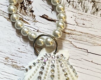 May include: A white seashell keychain adorned with small pearls and sparkling rhinestones. The shell is attached to a silver keyring and a loop of faux pearls. The keychain has a nautical theme.