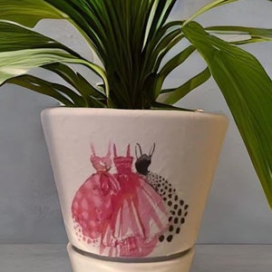 May include: A light pink ceramic plant pot with a flared shape, featuring a watercolor design of three dresses in shades of pink and black. The pot holds a lush green plant with large, textured leaves. The pot has a matching saucer.