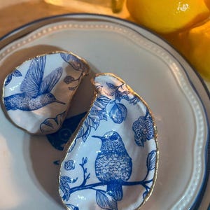 May include: Two white ceramic oyster shells with gold trim, each featuring a blue and white hummingbird design. The shells are on a white plate with a blue rim.