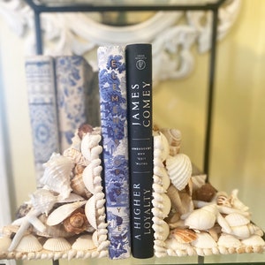 May include: A pair of bookends made of seashells, holding up two books. The book on the left is blue with gold lettering and the title "Elements of Family." The book on the right is black with white lettering and the title "A Higher Loyalty" by James Comey.