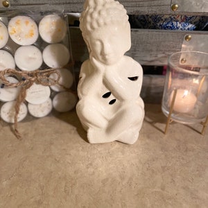 Puede incluir: Una estatua de Buda de cerámica blanca en posición meditativa. La estatua tiene tres pequeñas aberturas. También se ve un paquete de velas de té y una vela encendida en un portavelas de cristal.