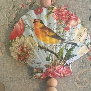 May include: A decorative seashell with a floral design and a yellow bird. The shell is painted with a blue background and features pink, red, and white flowers. The bird is perched on a branch with green leaves. The shell is hanging from a wooden bead garland.