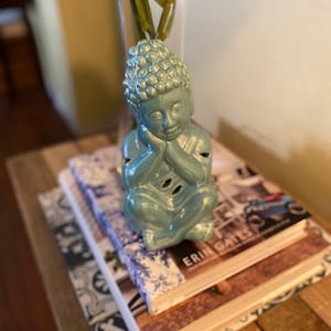 May include: A blue ceramic figurine of a meditating Buddha sitting on a stack of books. The top book has the text "Erin Gates" on the cover.