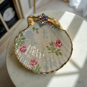 May include: A decorative seashell-shaped dish with a floral pattern of pink roses and blue leaves. The rim is trimmed with gold paint, and a jeweled embellishment is at the top. This unique dish is perfect for jewelry or small items.