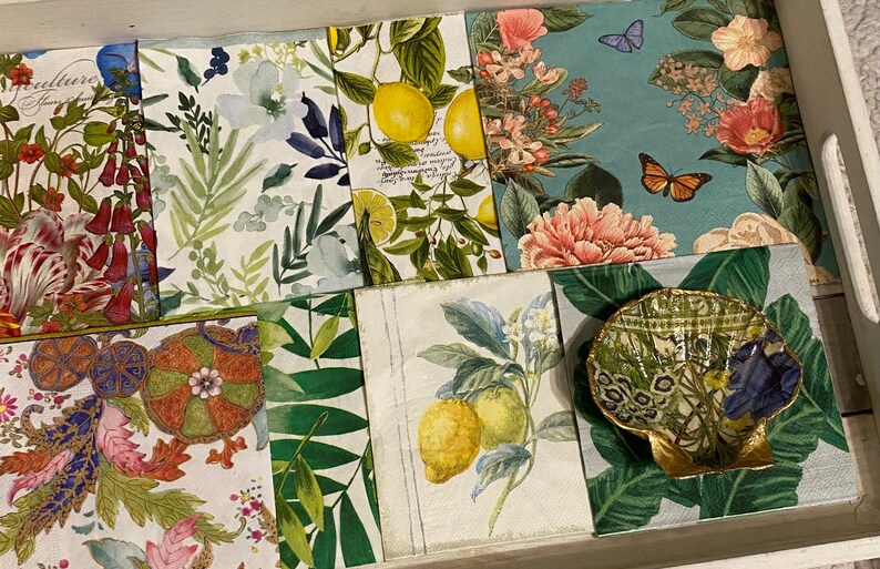 May include: A set of eight decorative paper napkins with floral and botanical designs. The napkins feature a variety of flowers, leaves, and fruits, including lemons, butterflies, and peonies. The napkins are arranged in a tray with a small decorative shell.