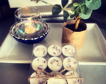 Op de afbeelding: Een transparante doos, vastgebonden met touw, met twaalf witte theelichtkaarsen met paarse accenten. De doos is gelabeld met "CANDLECHIC". Een kleine potplant en een decoratieve kom zijn ook zichtbaar.