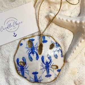 May include: A white ceramic sand dollar ornament with a gold trim and a blue and white lobster pattern. The ornament is hanging from a twine string.