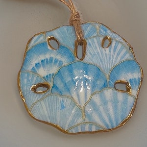 May include: A round, light blue and white sand dollar ornament with a gold-colored trim and a natural twine loop for hanging. The sand dollar design features a fan-like pattern. The ornament is a decorative item.