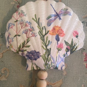May include: A white seashell with a floral and dragonfly design. The seashell is hanging from a string of wooden beads.