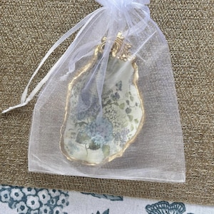 May include: A decorative oyster shell dish featuring a floral decoupage pattern with blue and green hydrangea blossoms, finished with a metallic gold painted edge and a small gold-toned metal embellishment at the top, enclosed in a white organza bag.