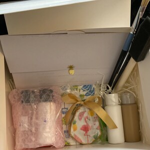 May include: A white box filled with craft supplies for a paint-your-own-ceramic project. The box contains two small bottles of paint, a ceramic piece with a flamingo design, paintbrushes, and a gold ribbon.