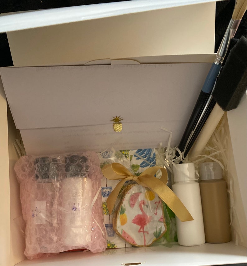 May include: A white box filled with craft supplies for a paint-your-own-ceramic project. The box contains two small bottles of paint, a ceramic piece with a flamingo design, paintbrushes, and a gold ribbon.