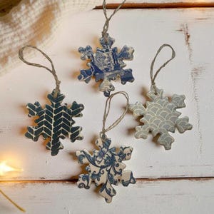 May include: Four snowflake-shaped ornaments with twine hangers. Each ornament has a unique design in shades of blue and white. The designs include a chevron pattern, floral motifs, and a wave-like pattern. These ornaments are perfect for holiday decorating.