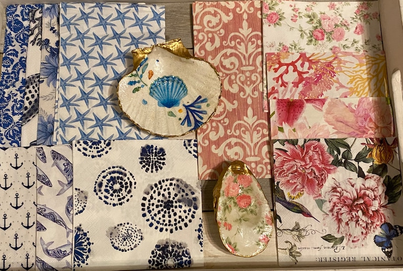 May include: A collection of decorative napkins with various patterns, including blue starfish, anchors, fish, and circles. Two decorative seashell dishes are included, one with a blue and white floral design and the other with a pink floral design.