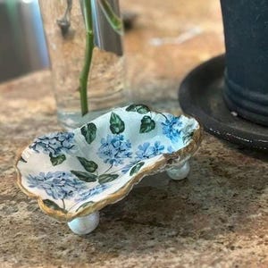 May include: A decorative, hand-painted oyster shell dish. The shell is white with blue hydrangea floral designs and green leaves. The rim is trimmed with gold paint. The dish is elevated on three small, white, pearl-like feet.