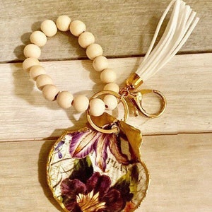 May include: A wooden bead keychain with a white tassel and a floral patterned seashell charm. The keychain is attached to a gold key ring.