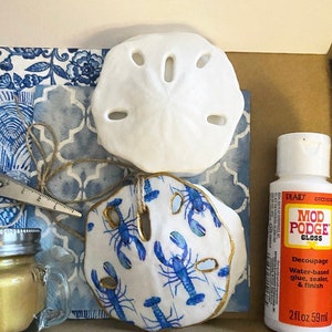 May include: A craft kit for making a nautical-themed sand dollar ornament. The kit includes a white sand dollar, a sand dollar with a blue and white lobster pattern, a small jar of gold paint, a bottle of Mod Podge gloss decoupage glue, and a piece of blue and white patterned fabric.