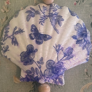 May include: A white seashell with a blue and white floral pattern and a butterfly design. The seashell is hanging from a wooden bead garland.