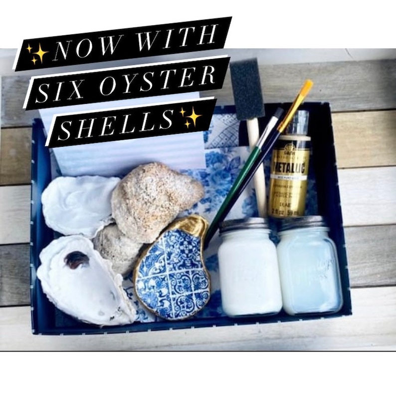 May include: A blue and white tray with six oyster shells, two jars of white paint, a gold paint bottle, and two paintbrushes. The text "NOW WITH SIX OYSTER SHELLS" is written on a white card in the tray.
