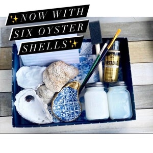 May include: A blue and white tray with six oyster shells, two jars of white paint, a gold paint bottle, and two paintbrushes. The text "NOW WITH SIX OYSTER SHELLS" is written on a white card in the tray.