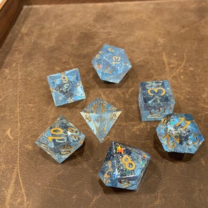 May include: A set of seven blue dice with gold numbers and glitter. The dice are on a brown leather surface.