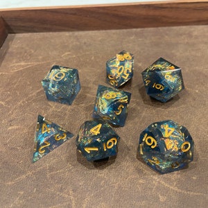May include: A set of seven blue and gold dice with a galaxy-like pattern. The dice are a d4, d6, d8, d10, d%, d12, and d20.