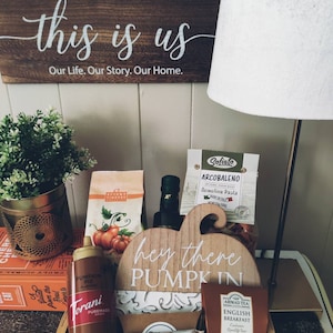 May include: A wooden gift basket filled with fall-themed treats, including a pumpkin pie spice, a bottle of olive oil, a box of pasta, a box of tea, and a wooden sign that says "hey there pumpkin".