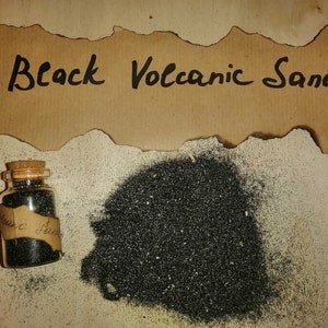 Op de afbeelding: Een klein glazen potje met een kurken deksel gevuld met zwart vulkanisch zand. Het potje staat op een bruine papieren achtergrond met de tekst "Black Volcanic Sand" erop.
