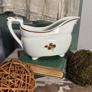 May include: White ceramic gravy boat with a brown floral design and trim. The gravy boat has a handle and sits on three small feet. The boat is resting on top of a vintage book.