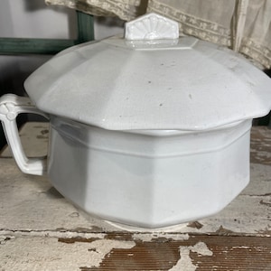 May include: A white, antique chamber pot with a lid. The pot has a handle and a geometric design. The lid features a decorative handle. The pot is sitting on a distressed, white-painted wooden surface.