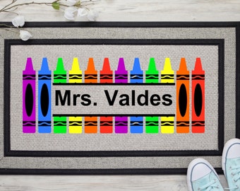 May include: A welcome mat with a gray background and black border. The mat features a row of colorful crayons with the text "Mrs. Valdes" in black.