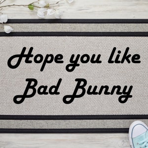 May include: A rectangular doormat with the text "Hope you like Bad Bunny" in black script. The mat has a light grey centre with a black border. A pair of light blue trainers are visible at the bottom.