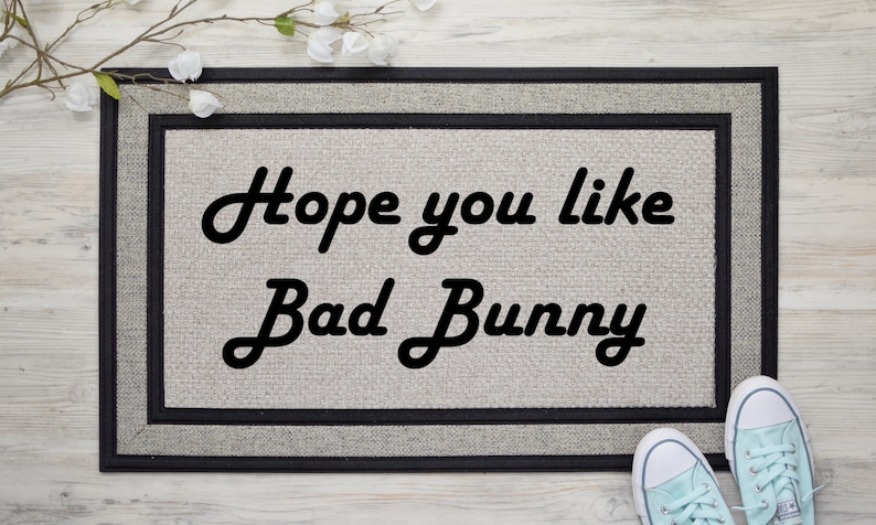 May include: A rectangular doormat with the text "Hope you like Bad Bunny" in black script. The mat has a light grey centre with a black border. A pair of light blue trainers are visible at the bottom.