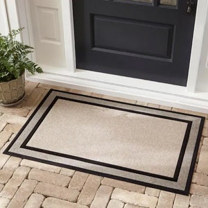 May include: A rectangular doormat with a neutral beige centre and a double border. The outer border is black, and the inner border is a lighter grey. The mat is placed on a brick patio in front of a black door.