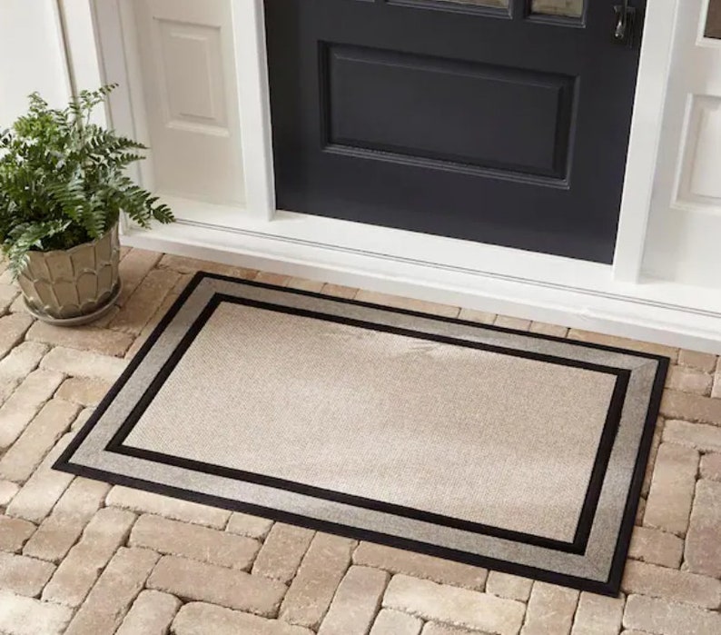 May include: A rectangular doormat with a neutral beige centre and a double border. The outer border is black, and the inner border is a lighter grey. The mat is placed on a brick patio in front of a black door.