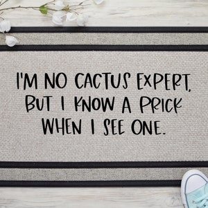 May include: A gray doormat with black trim and the text "I'M NO CACTUS EXPERT, BUT I KNOW A PRICK WHEN I SEE ONE."