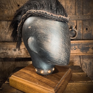 May include: A dark gray mannequin head with a black hairpiece and a wooden base. The hairpiece has a woven band with red and brown accents. The base is a square of polished wood. The head has a metallic finish.