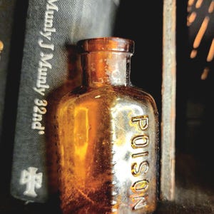 Könnte beinhalten: Kleine, antike Flasche aus bernsteinfarbenem Glas mit dem Wort „POISON“ auf der Vorderseite. Die Flasche ist zylindrisch mit abgerundeter Schulter und kurzem Hals. Im Hintergrund ein Stapel Bücher.