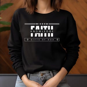 May include: Black sweatshirt with the words "WISDOM FAITH KNOWLEDGE FAITH GIFTS OF GOD" in white block letters. The sweatshirt has a crew neck and long sleeves. The model is wearing blue jeans.