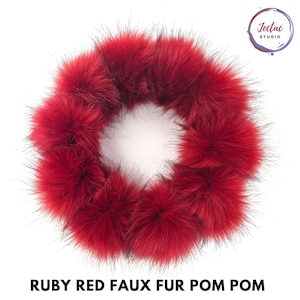 May include: A circular arrangement of ruby red faux fur pom poms. The fluffy, textured spheres are connected to form a ring shape. The text Jectac STUDIO appears in the top right corner, and the text RUBY RED FAUX FUR POM POM is at the bottom.