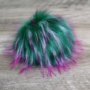 May include: A fluffy, round pompom in shades of green, white, and magenta. The faux fur has long, textured strands, creating a soft, voluminous appearance. The color combination is vibrant and eye-catching, suitable for various craft projects.
