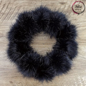 May include: A black faux fur pom pom trim, made of fluffy, soft, and plush material. The trim is arranged in a circle, creating a decorative wreath-like shape.
